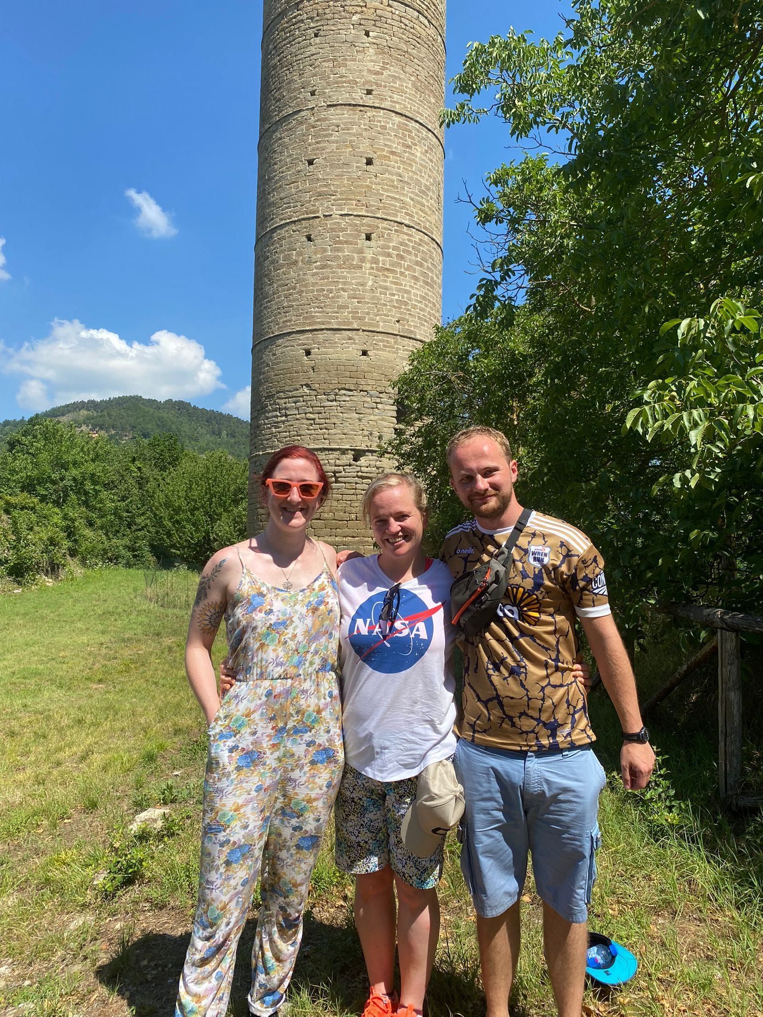 Three people pose in front of a tall, stone tower on a sunny day.