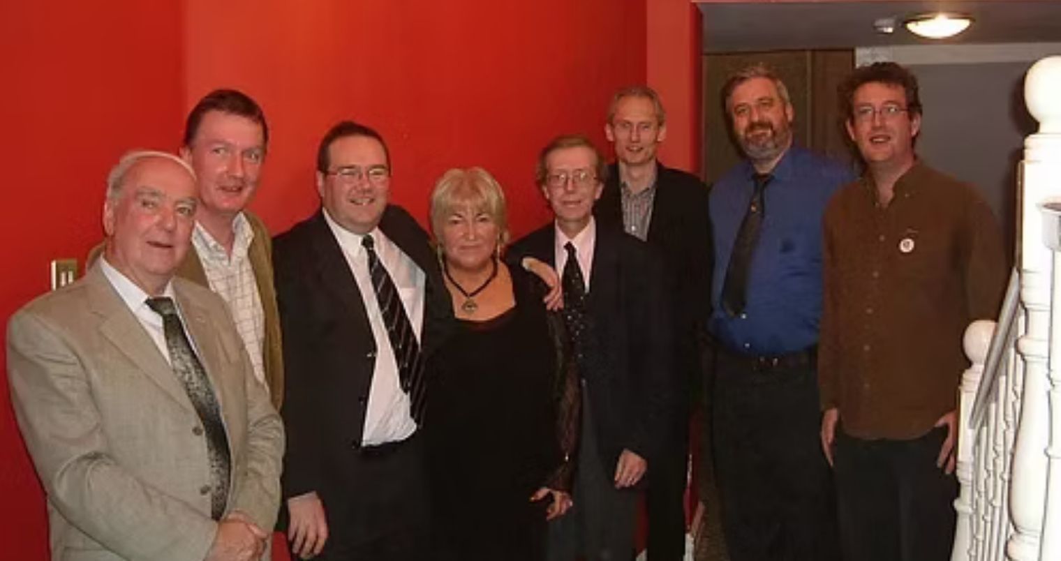 Group of people posing in front of a red wall. They are smiling, and some are wearing suits. A staircase is visible.