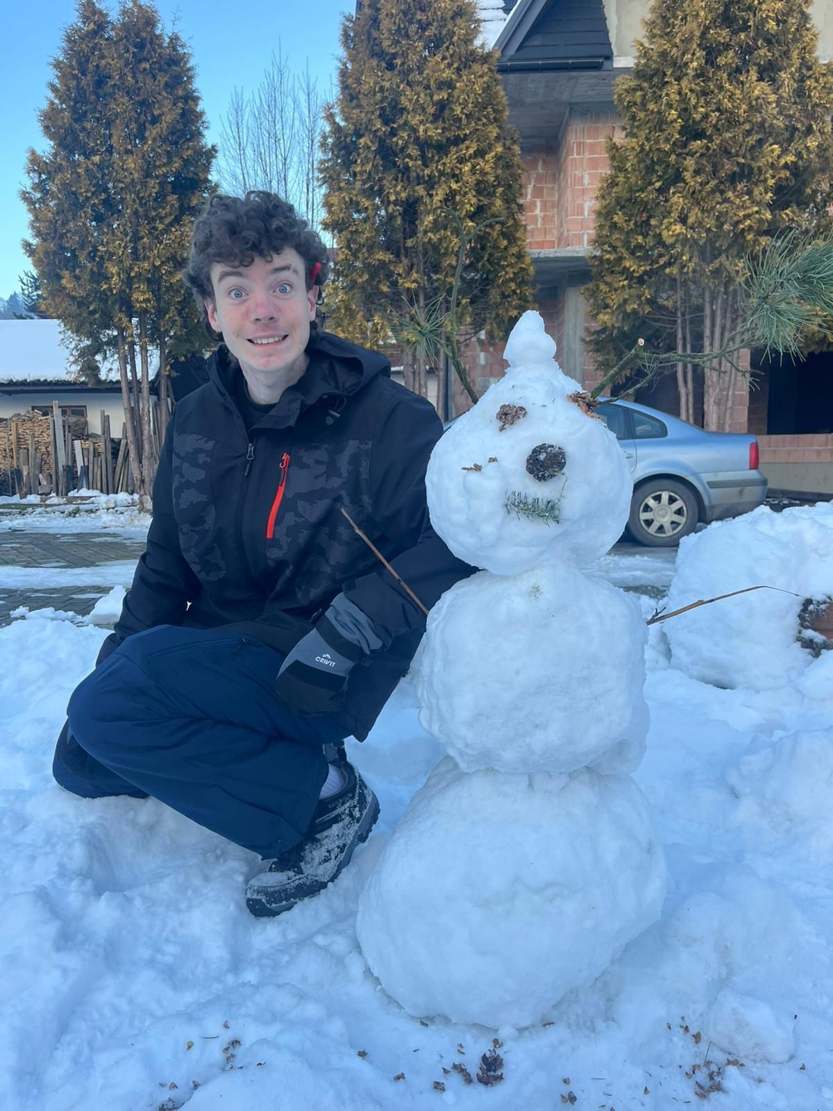 Person kneels next to a snowman in snow. The person smiles, wearing a jacket and dark pants. The setting is outdoors.