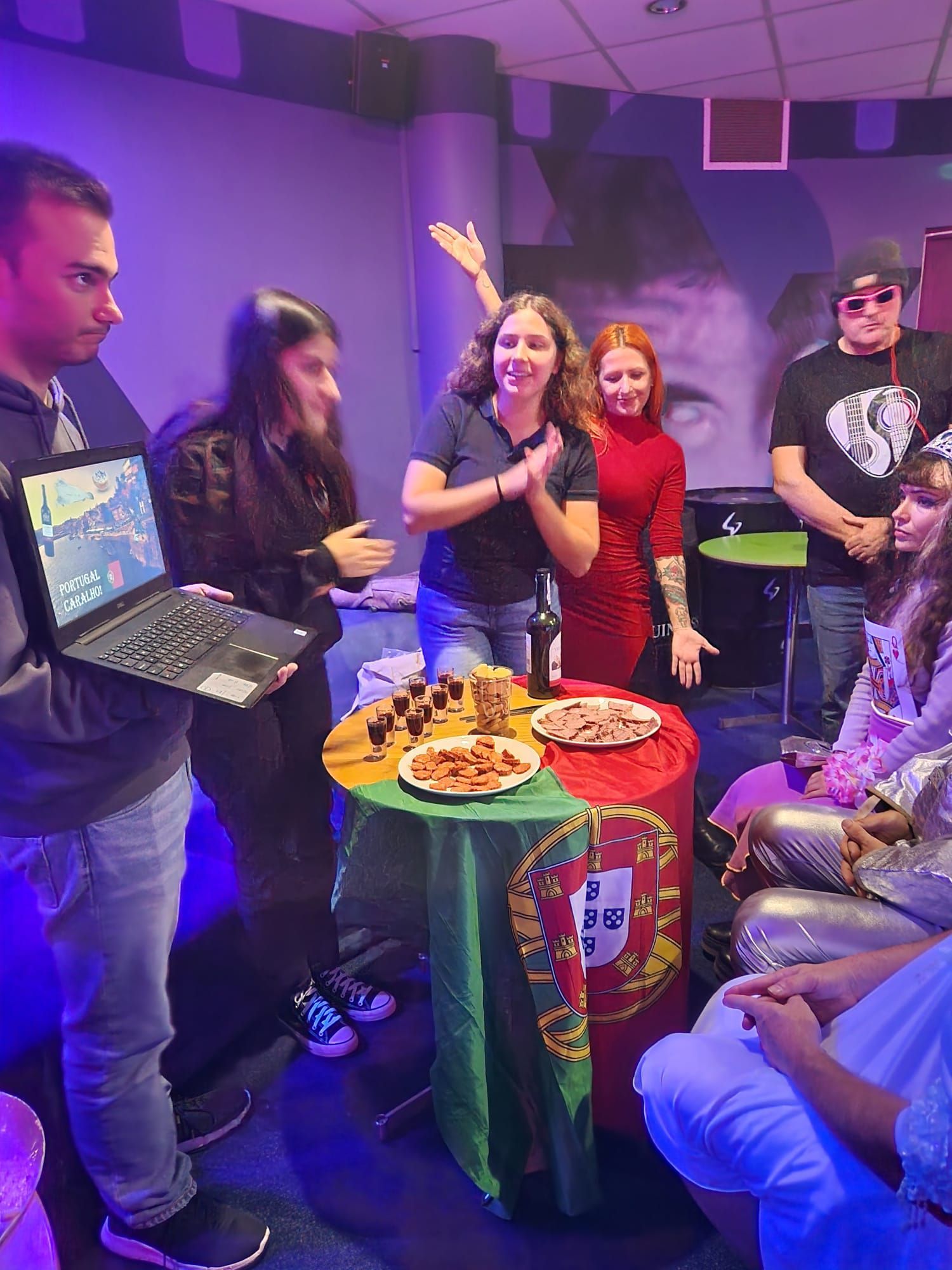 Group celebrates around table with Portuguese flag. Food and drinks are present. Cheerful expressions.