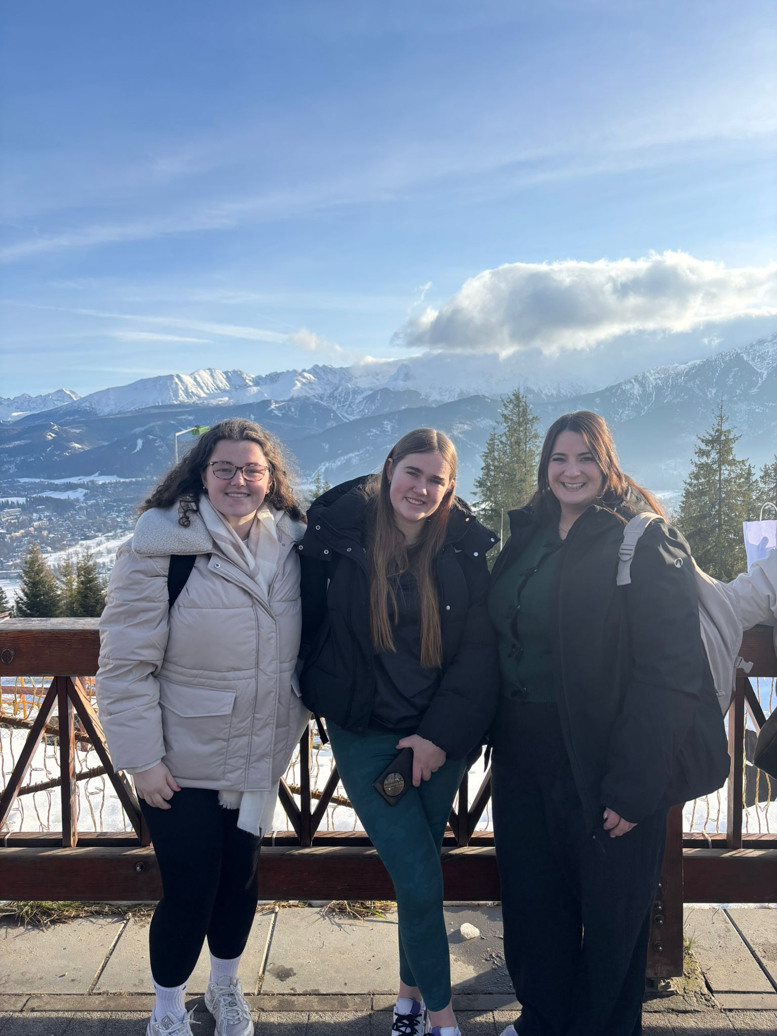 Three people pose outdoors with snow-covered mountains in the background. Bright sunlight, blue sky.
