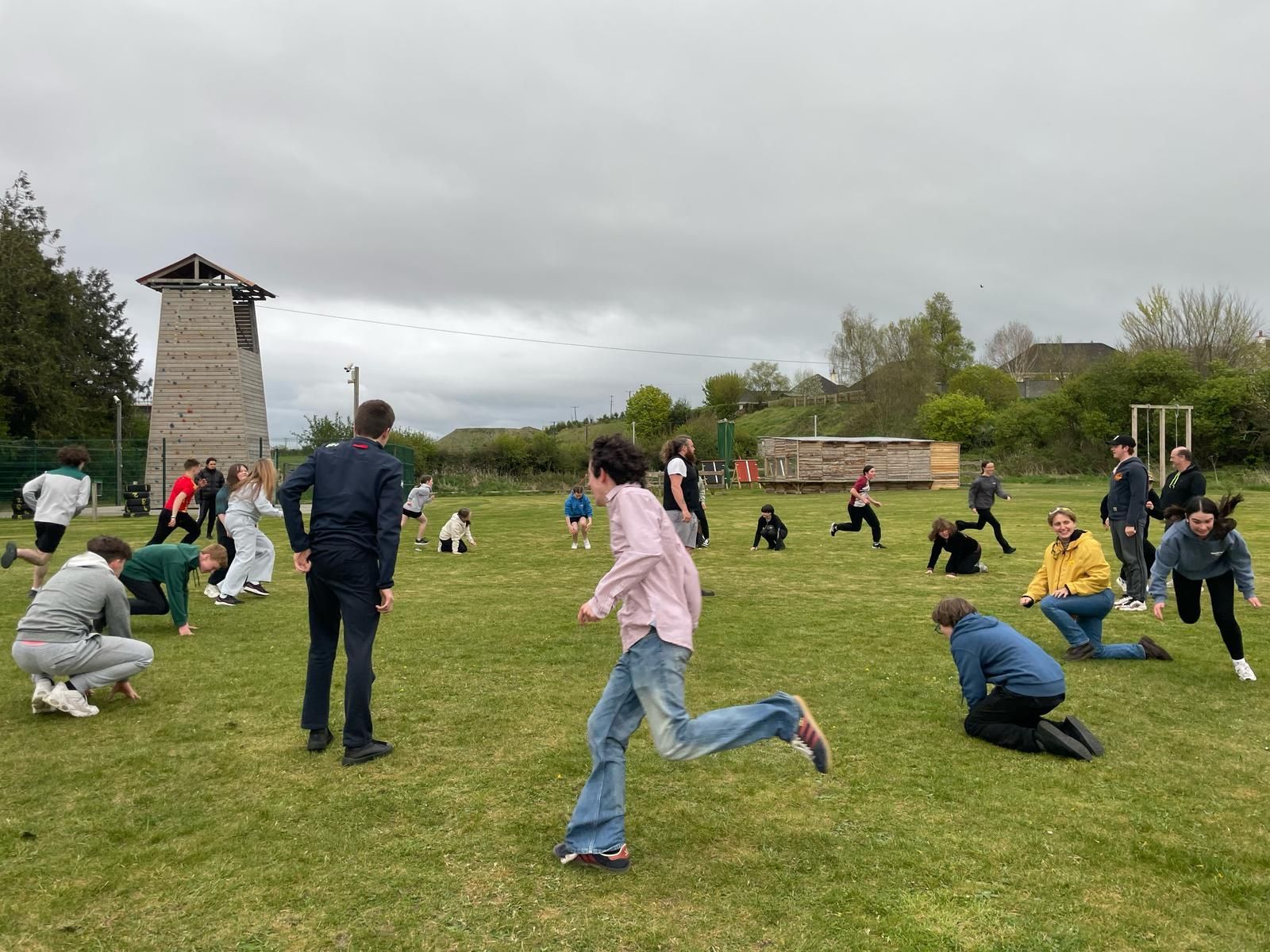 People running and exercising on a grassy field with a wooden tower and cloudy sky.