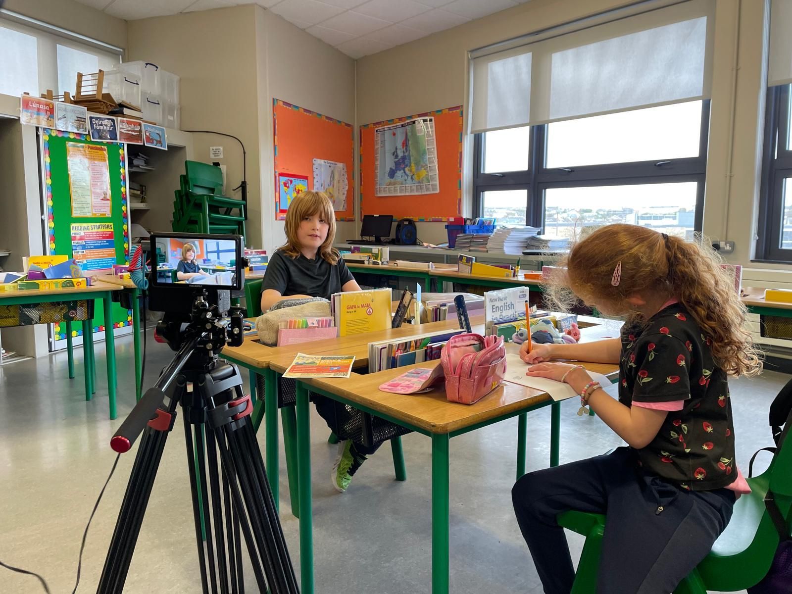 Two children at a desk recording a video in a classroom, one child writing.