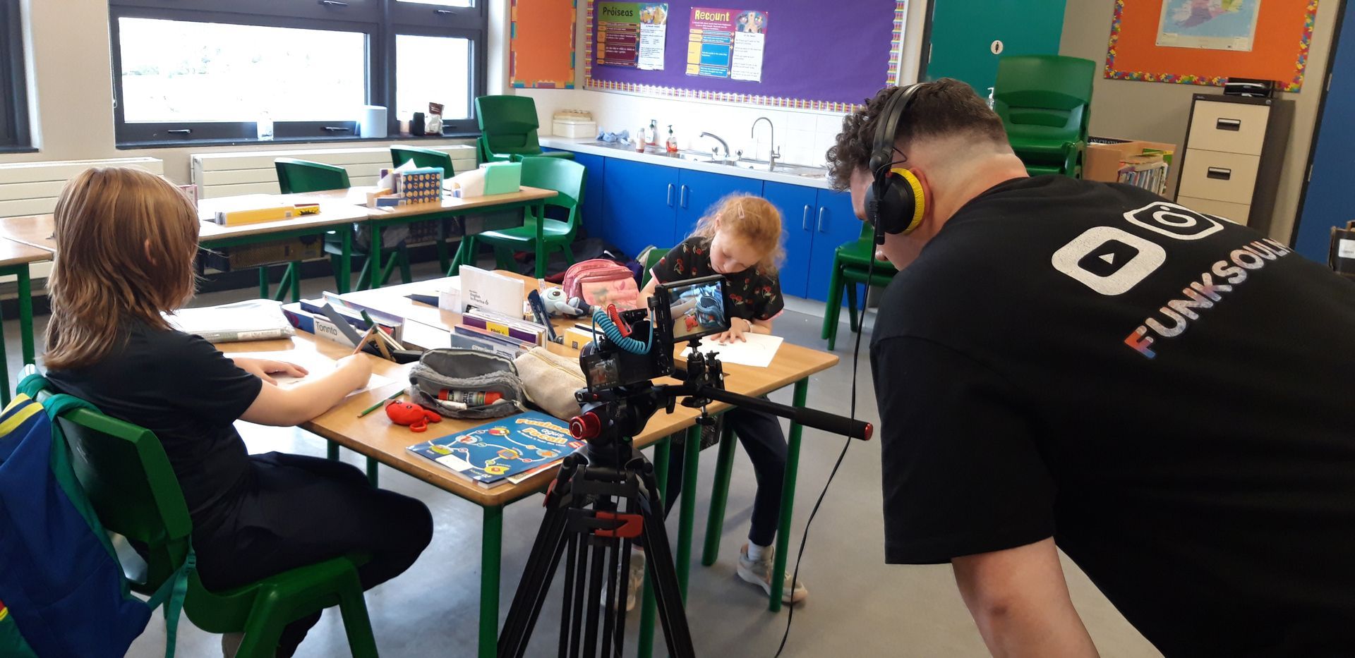 A person filming two children at desks in a classroom. The children are drawing.