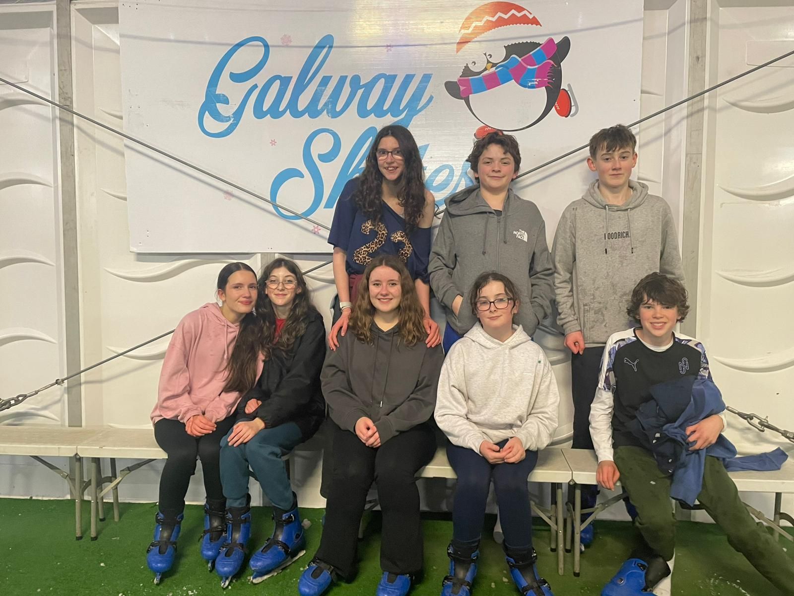 Group of people pose for photo at Galway Skates. They wear ice skates and stand near sign.