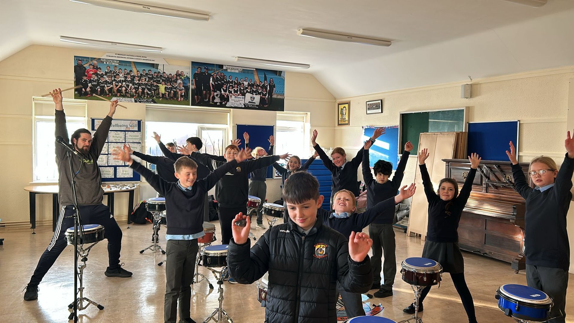 Group of students in a room, raising drumsticks. Teacher watches. Posters and drums visible.