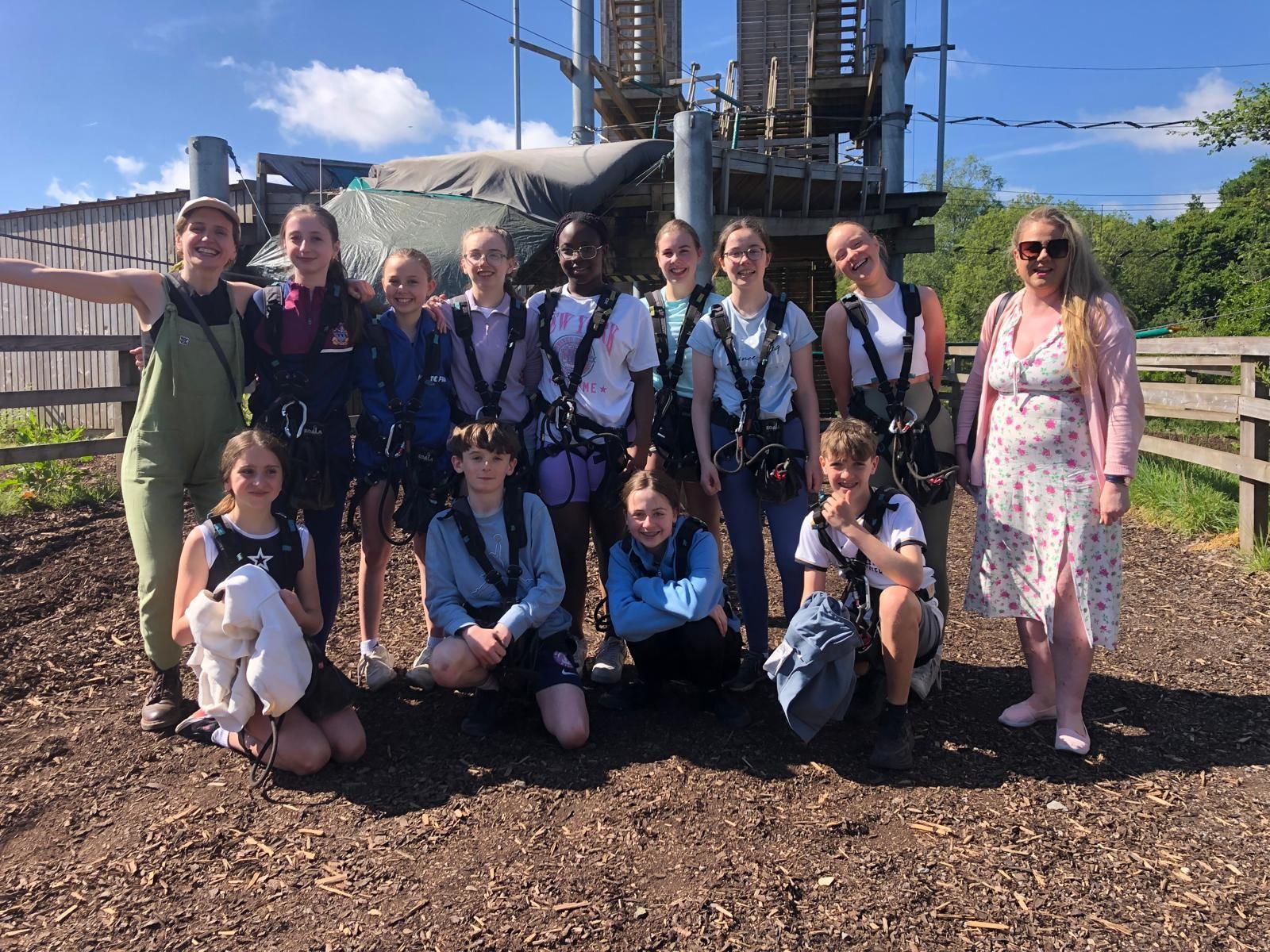 Group of people wearing harnesses in front of an adventure course; brown ground, sunny day.