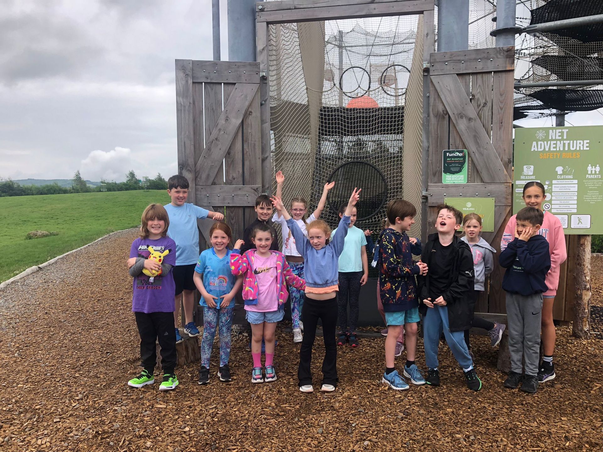 Group of children smiling and raising their arms in front of wooden play structure at an outdoor adventure park.