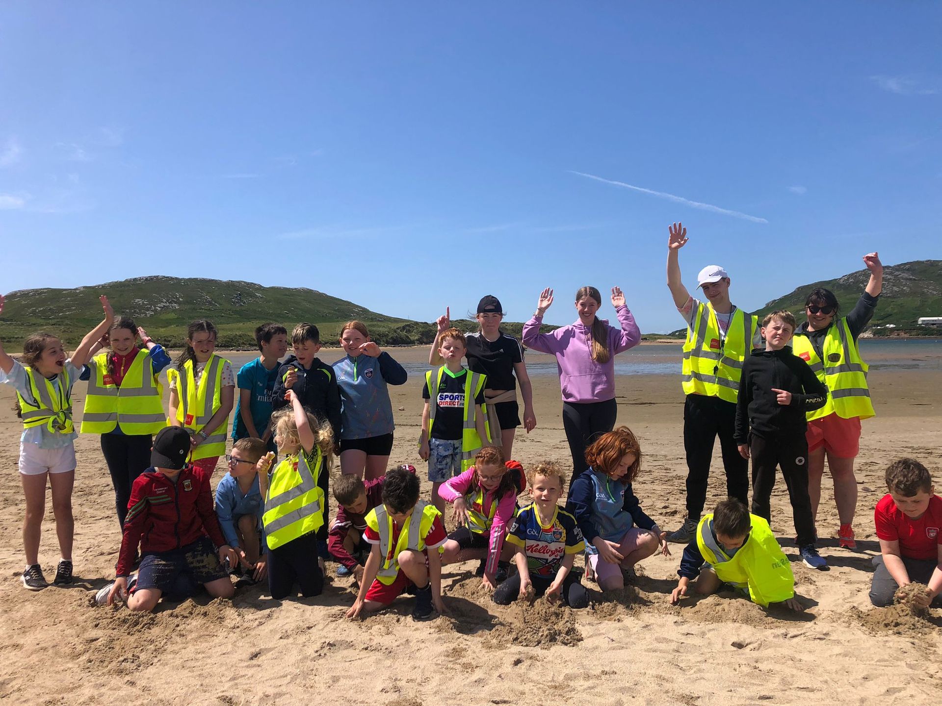 Group of people on a beach. Some wear yellow vests, some raise their arms. Blue sky, water, and hills in the background.