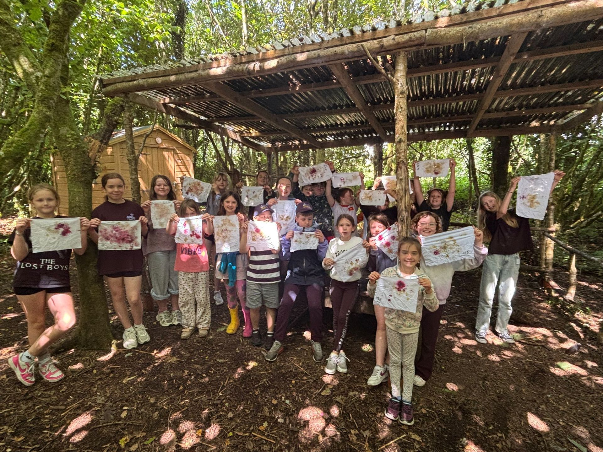 Group of children holding up artwork under a wooden shelter in a forest.