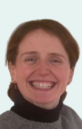 Woman with short brown hair smiles, wearing a black turtleneck and gray sweater.