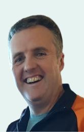 Man with short graying hair, smiling broadly; wearing a dark and orange zip-up top.