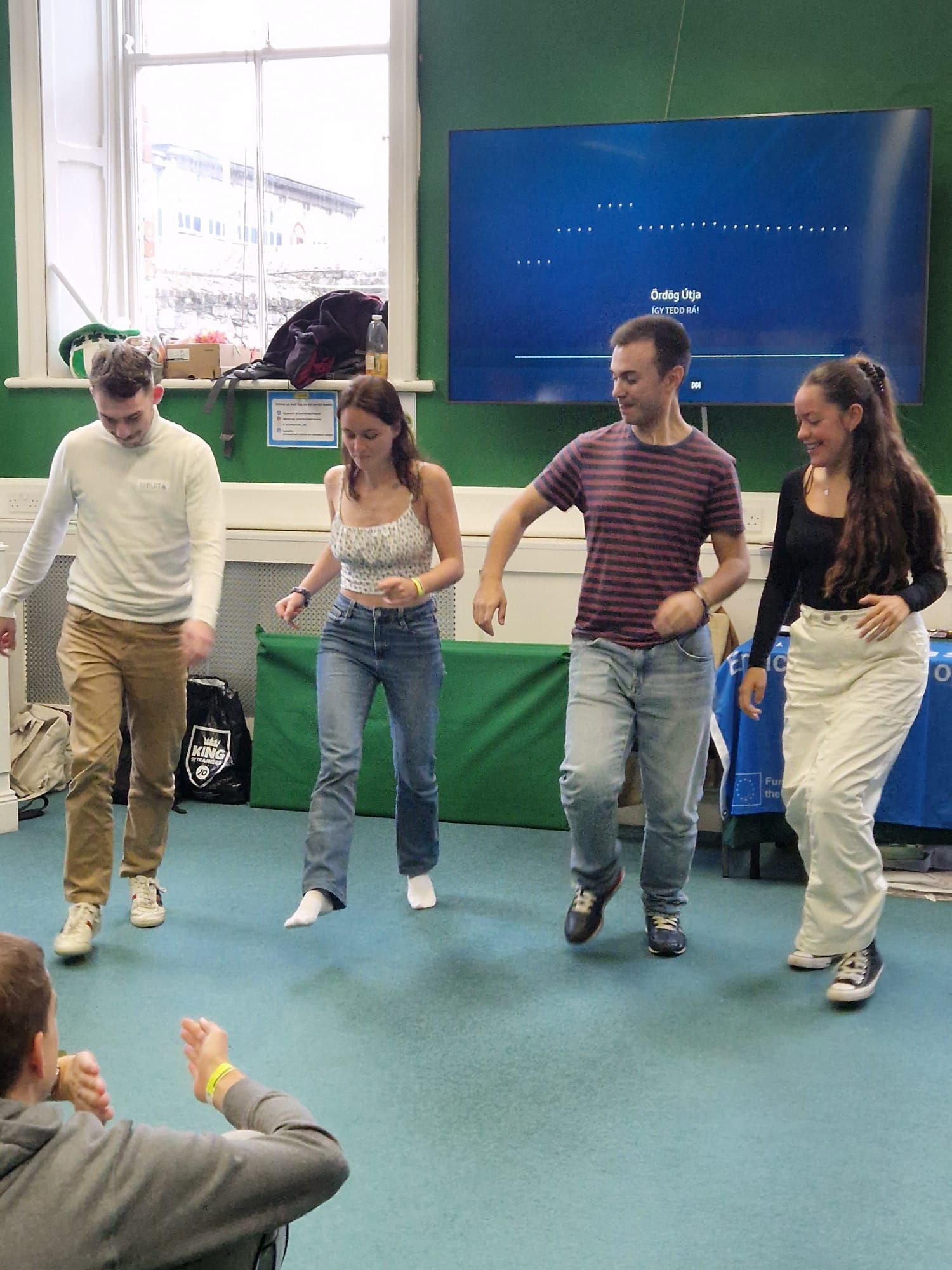 Four people dancing in a room. One person watches from foreground. Light coming from a window.