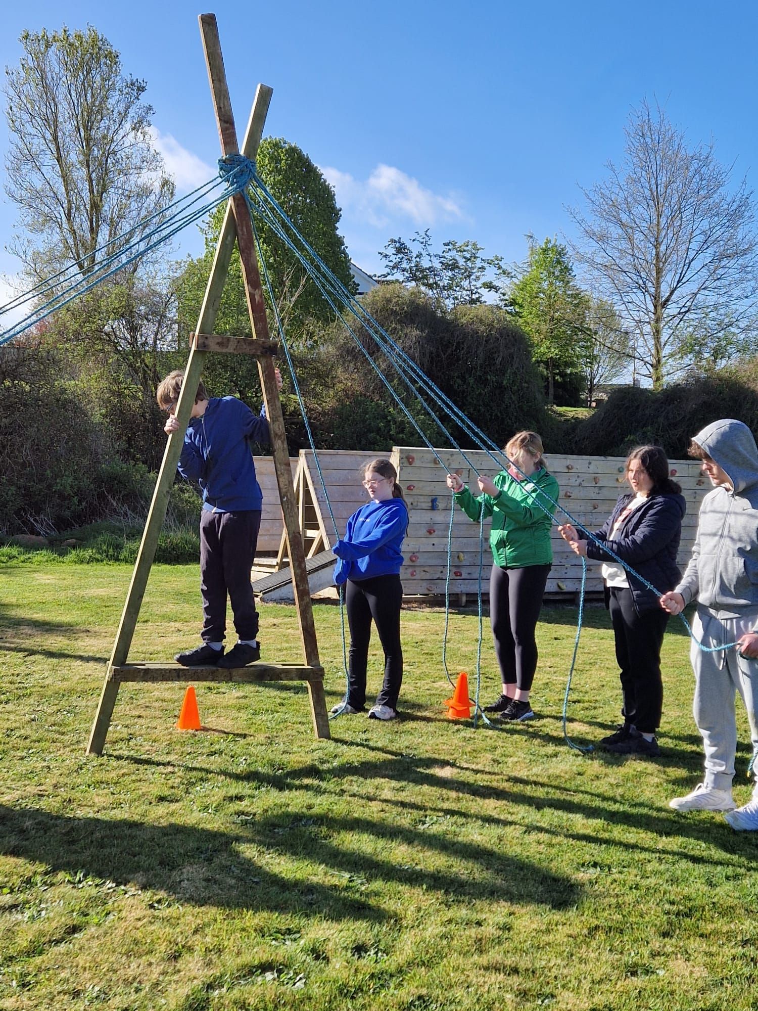 Group doing an outdoor teamwork challenge: wooden structure with ropes. One person climbs; others hold poles. Sunny day.