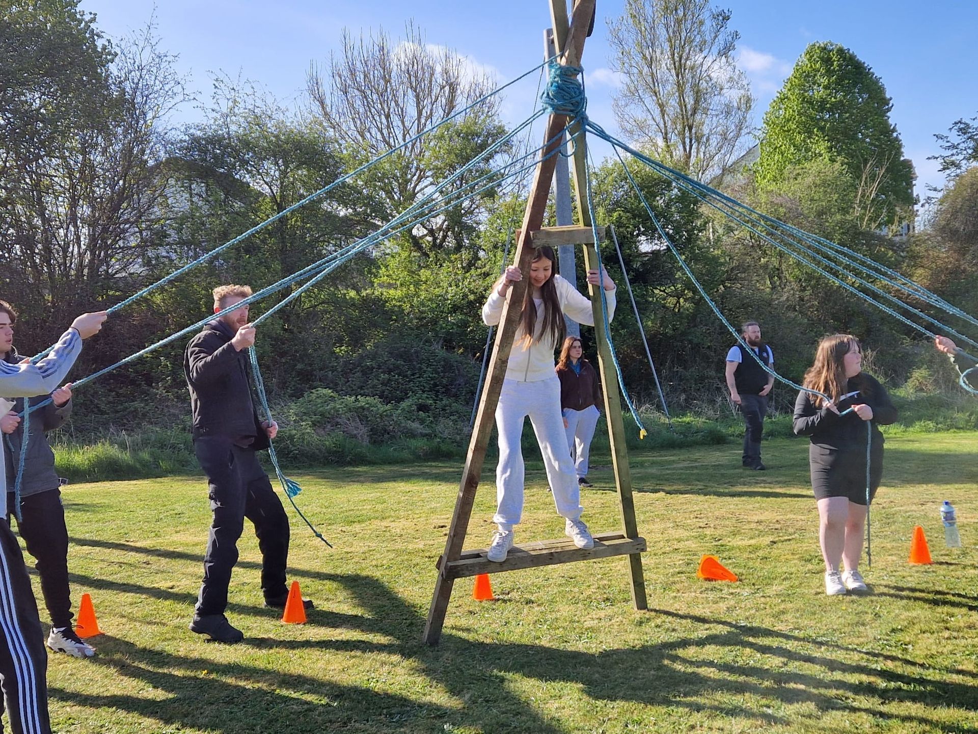 People participate in an outdoor team-building exercise, using ropes and a wooden structure.