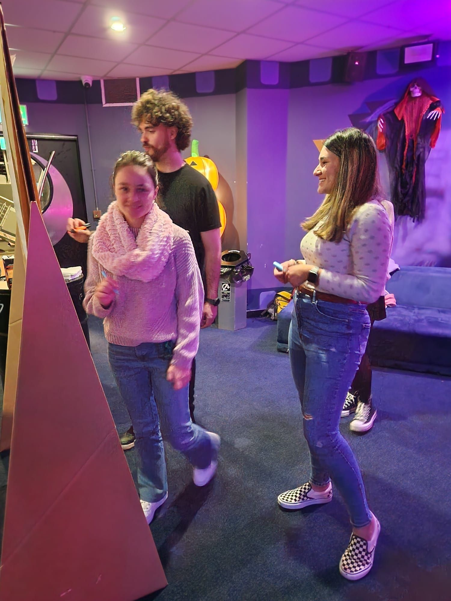 Three people in a room with arcade game decor. One woman points at a game, another woman smiles, and a man looks on.