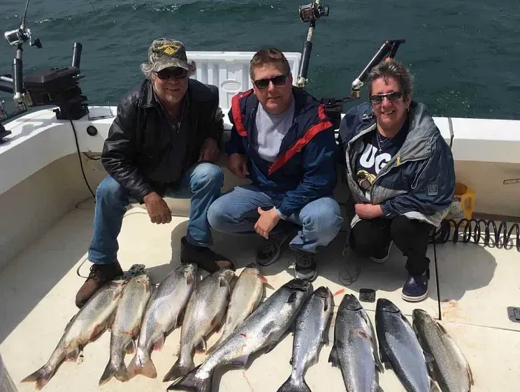 Three people are sitting on a boat with a bunch of fish on the deck.
