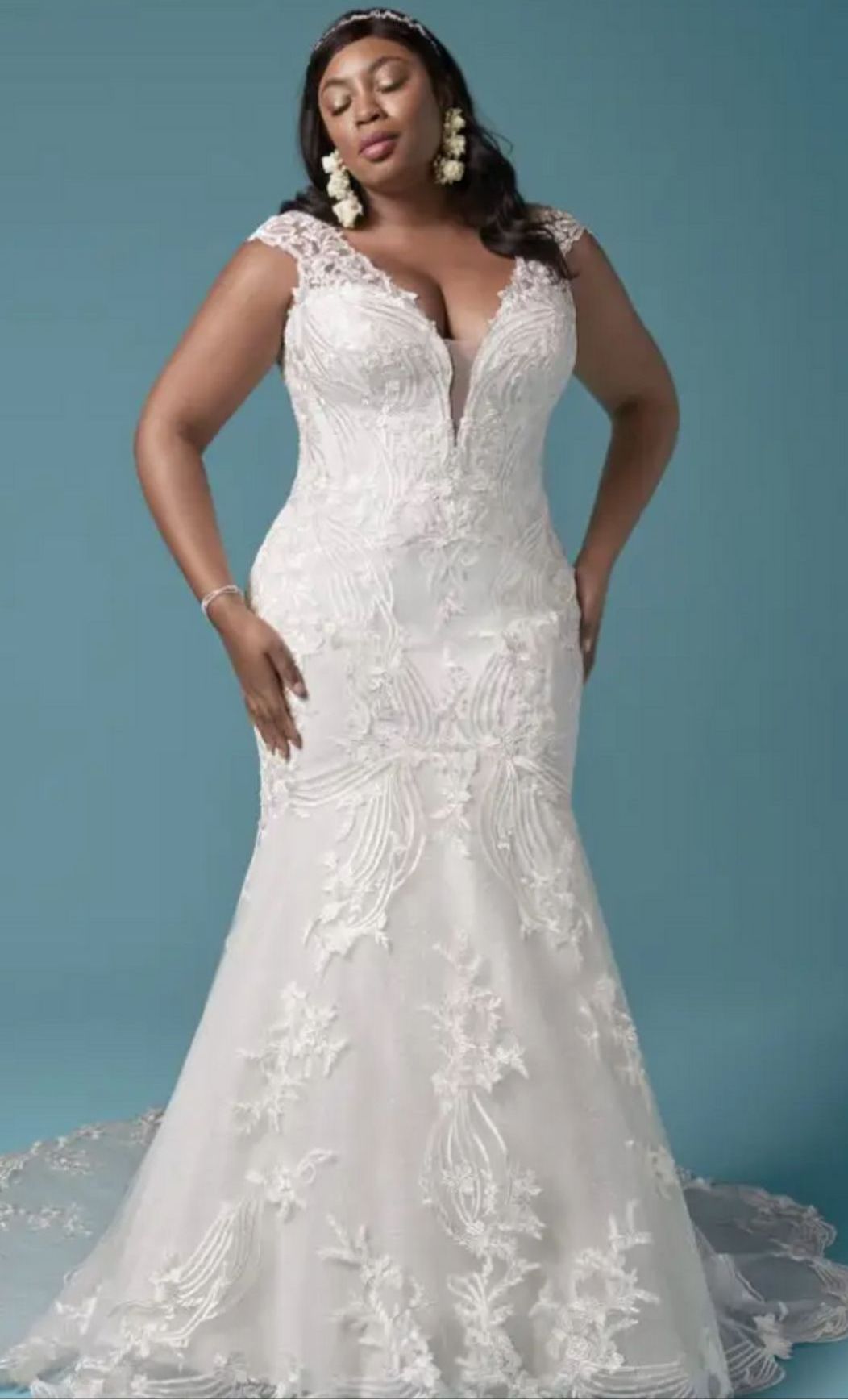 Woman in white lace wedding dress with a deep v-neckline and mermaid silhouette, standing in front of a blue background.