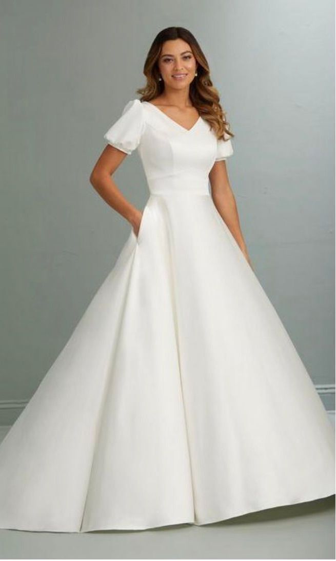 Woman wearing a white wedding dress with short sleeves and a full skirt. She is smiling, standing indoors.