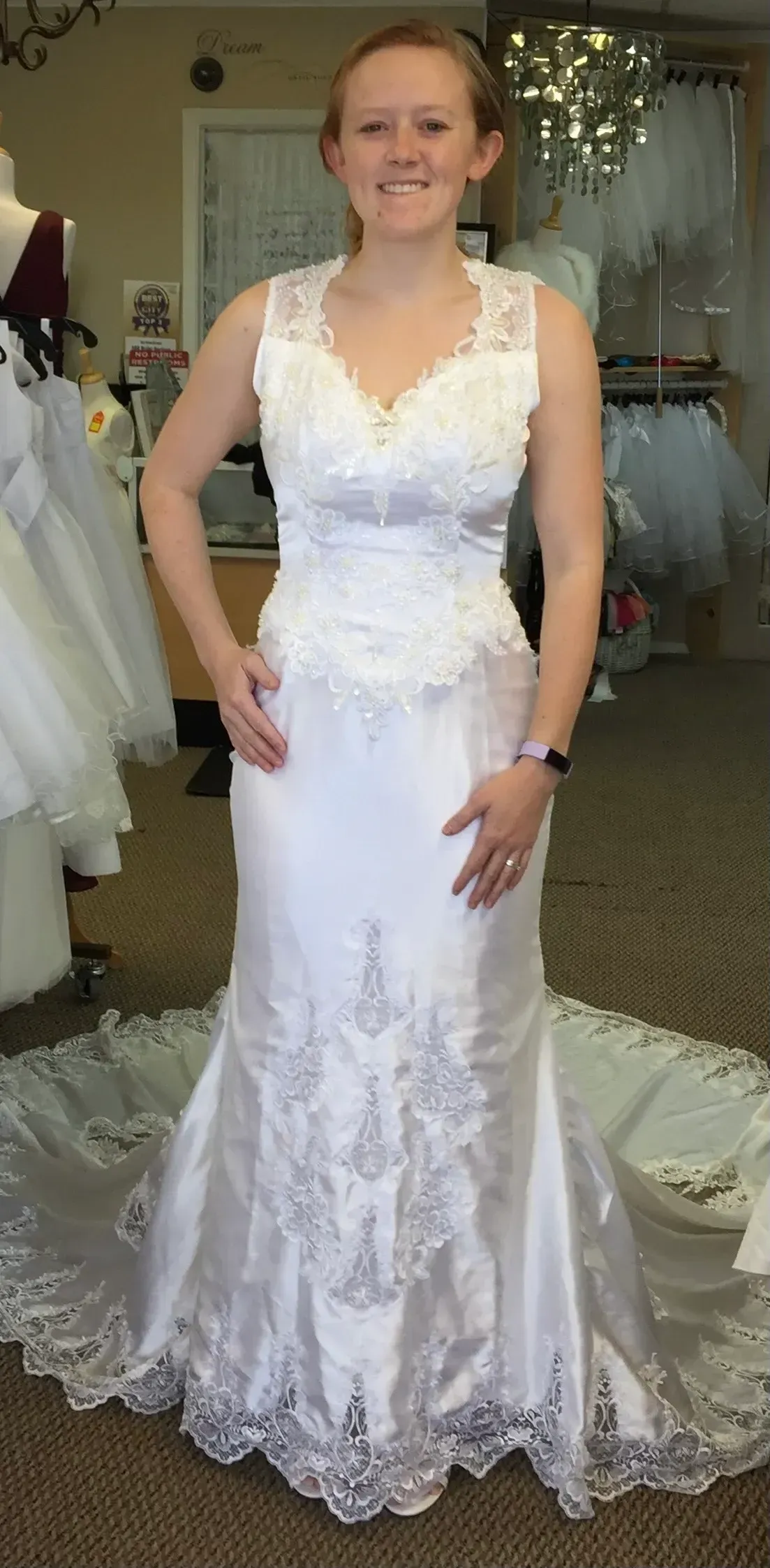 Woman wearing a white sleeveless wedding dress with lace detail, smiling, in a bridal shop.