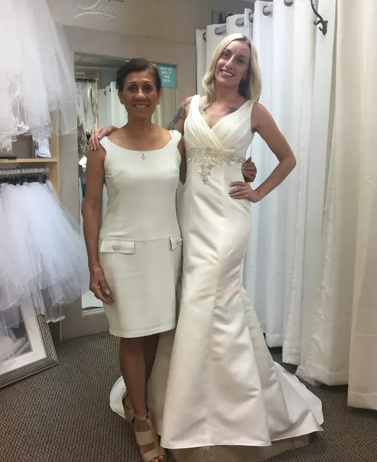 Woman in a white wedding dress with another woman. Both are in a bridal shop.