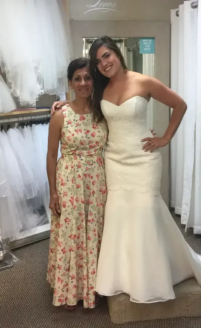Woman in a wedding dress stands with woman in floral dress inside a bridal shop.