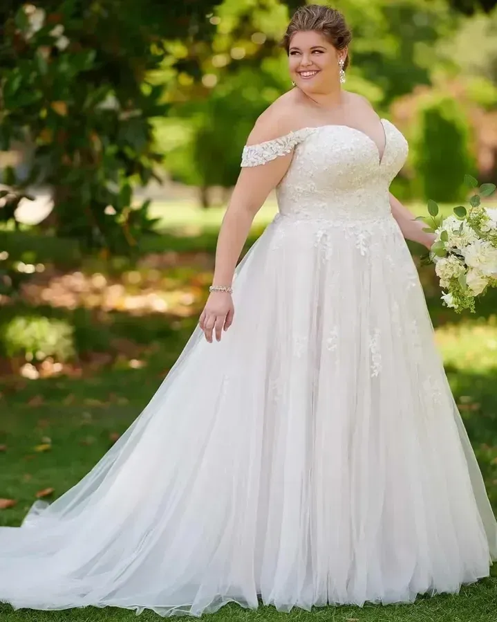 Woman in a white off-the-shoulder wedding gown holding a bouquet outdoors.