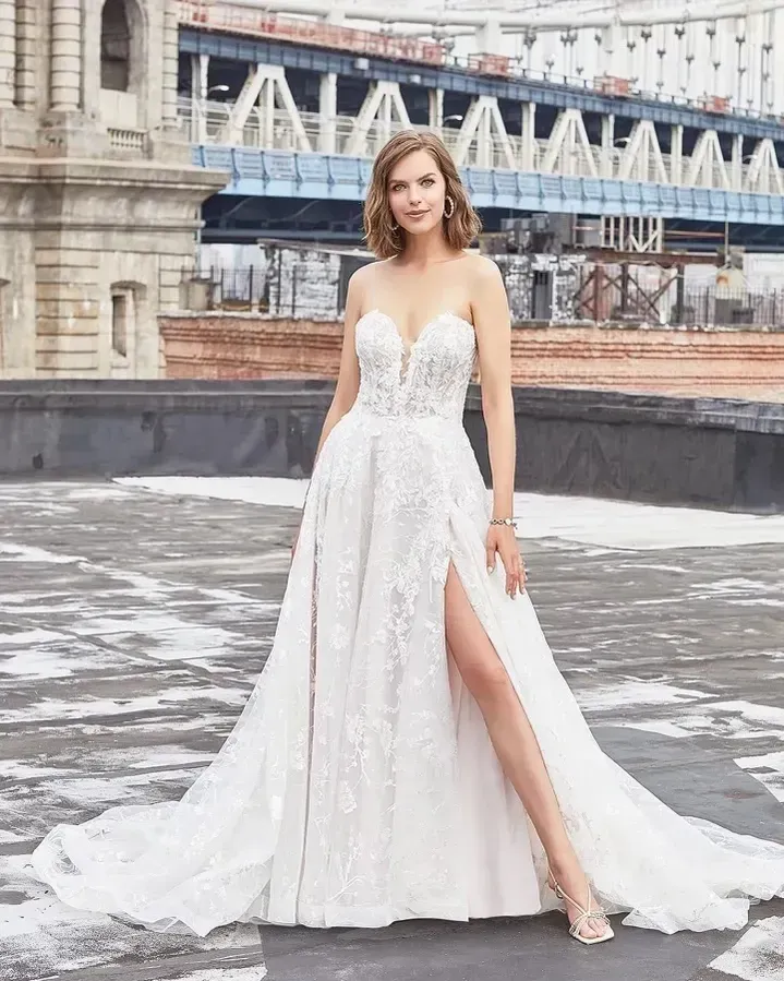Woman in strapless white wedding dress with high slit, standing on a rooftop, Manhattan Bridge in background.