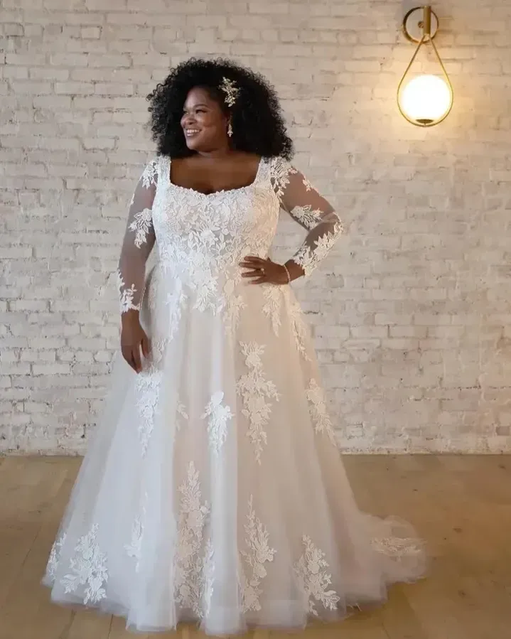 Woman in a white wedding gown smiles, standing against a white brick wall.