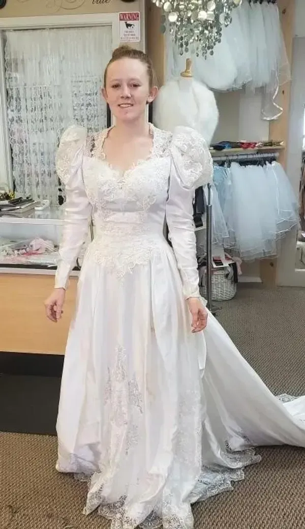 Woman in a white wedding dress with long sleeves and a train, smiling in a shop.