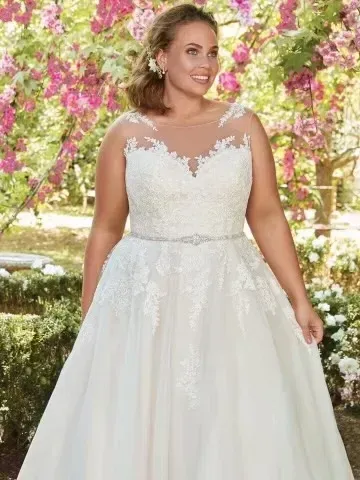Woman in a wedding dress, smiling, outdoors. She is wearing a sleeveless dress with a jeweled belt and sheer neckline.