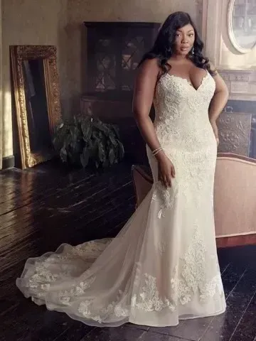 Woman in a white lace wedding dress with a long train, standing in a room with a mirror and fireplace.