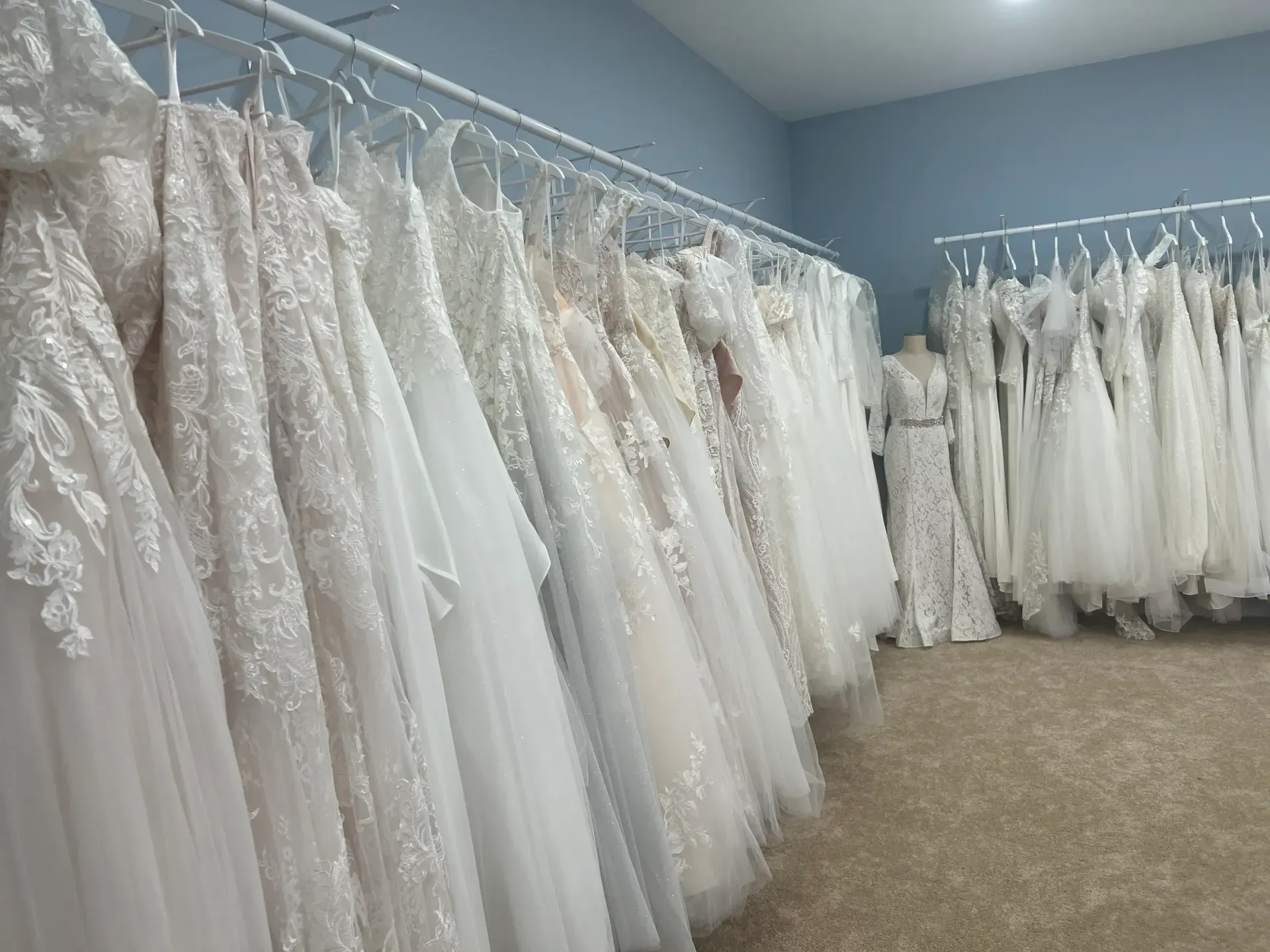 Wedding dresses hanging on racks in a bridal shop, soft lighting, and blue walls.