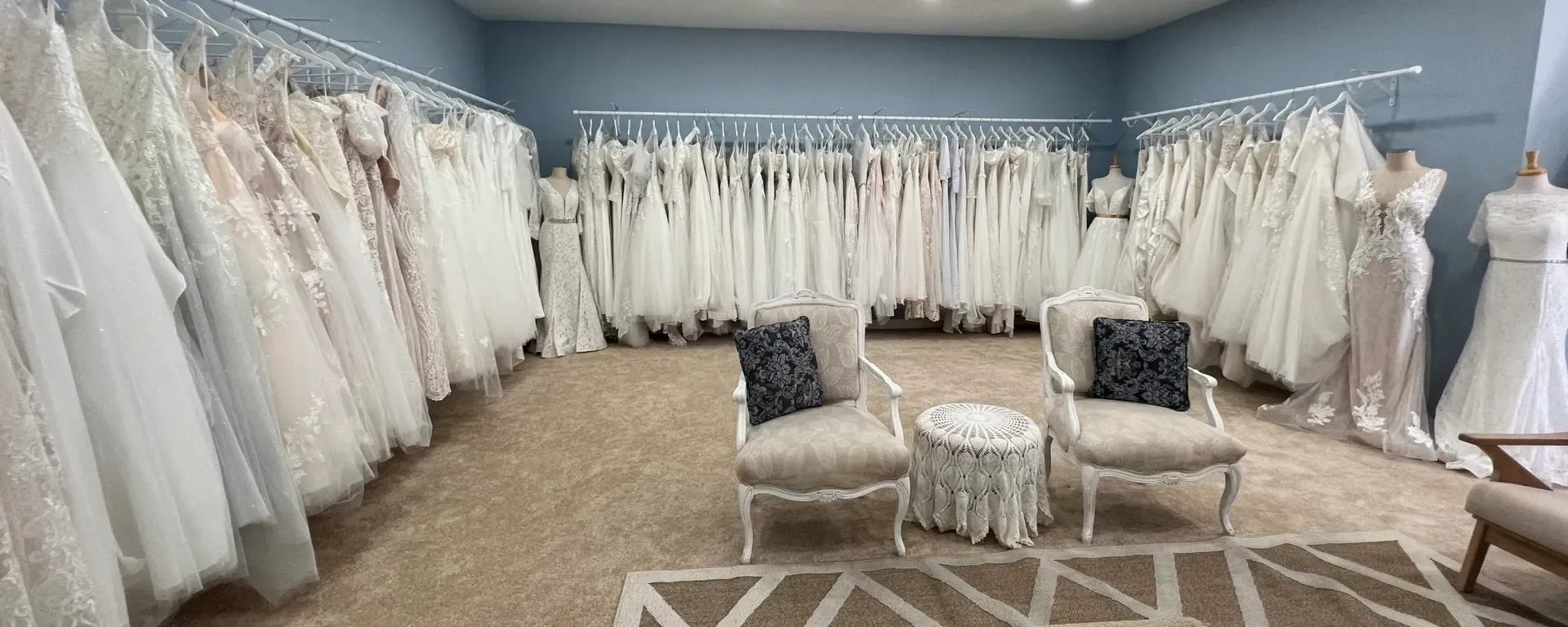 Wedding dresses displayed in a boutique. Dresses hang on racks; chairs and a small table are in the room.
