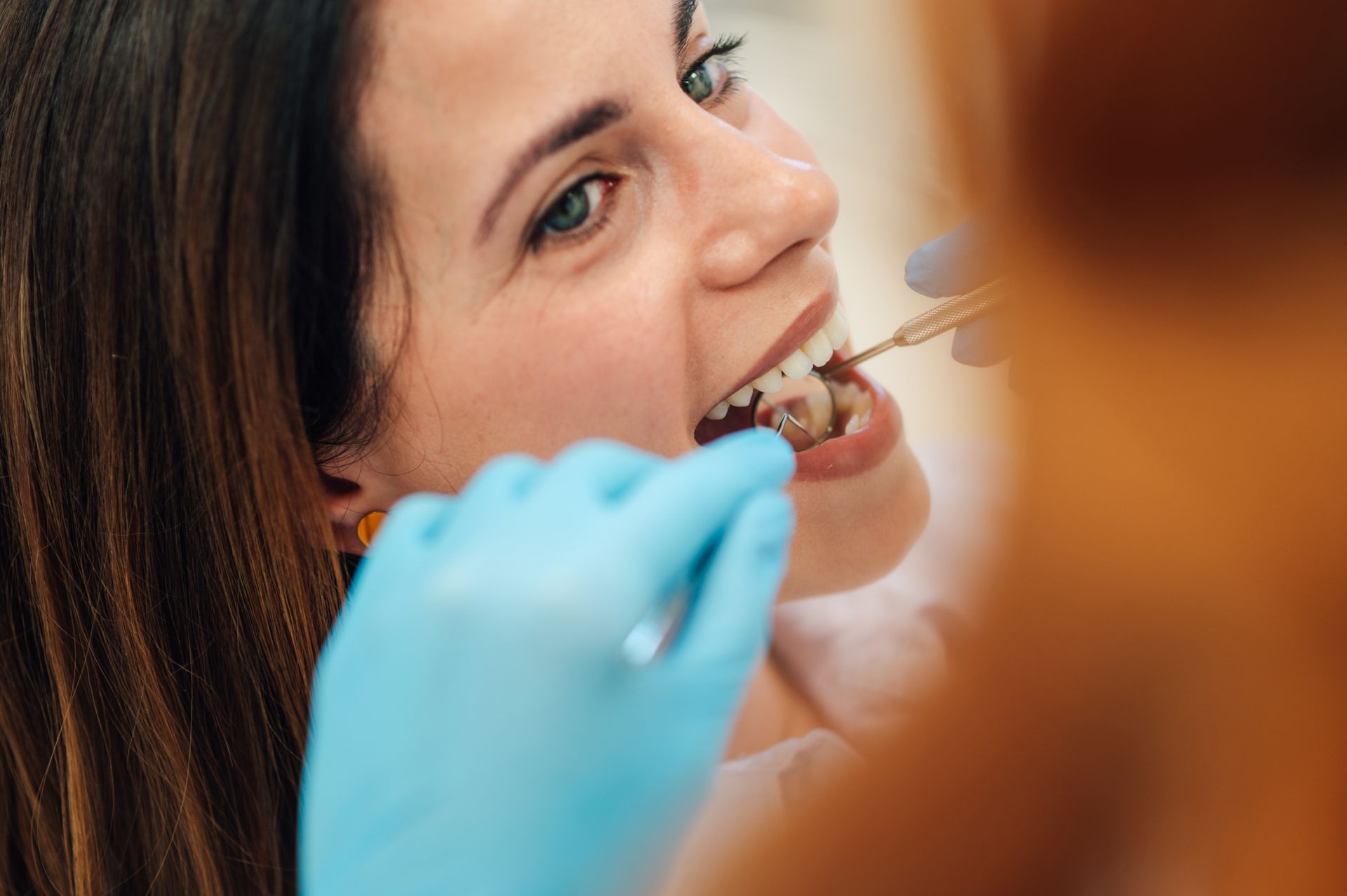 A dental professional in blue medical gloves uses a metal tool to examine a patient's teeth in a clinical setting.