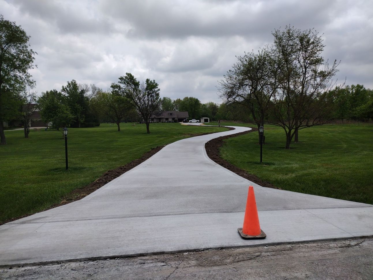Concrete driveway curving through a grassy yard under a cloudy sky, with a traffic cone on the edge.