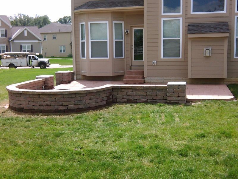 Brick patio and retaining walls adjacent to a beige house with a bay window, and a green lawn.