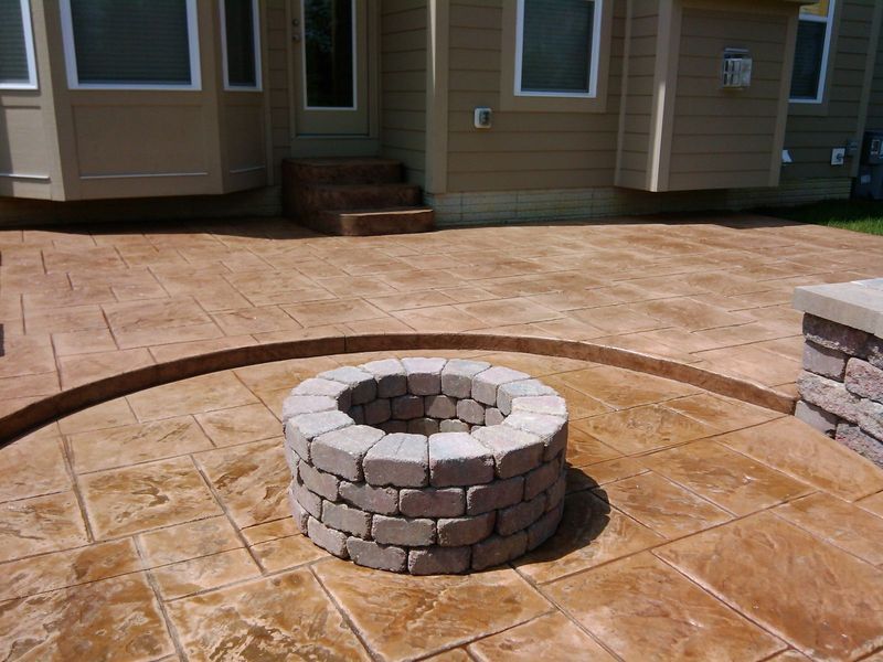 Stamped concrete patio with a circular fire pit and a beige house in the background.