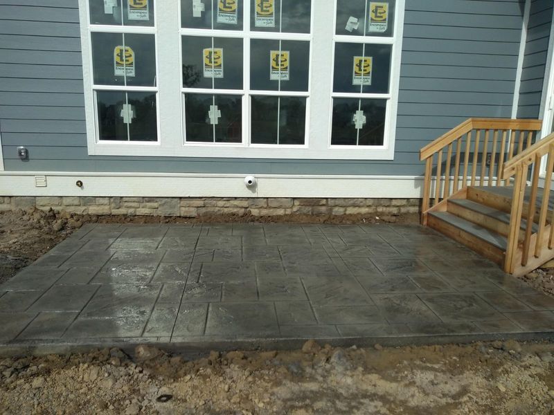 Concrete patio with textured surface, next to a building with a window, steps, and siding.