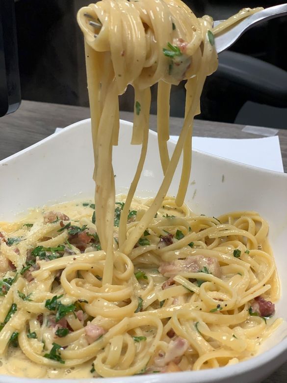 Fork lifting creamy spaghetti from a white bowl of pasta with herbs and bacon bits