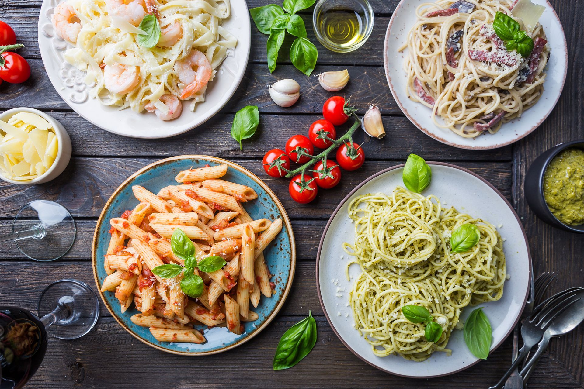 Four plates of pasta on a rustic table with tomatoes, herbs, garlic, and wine glasses. Four plates of pasta on a rustic table with tomatoes, herbs, garlic, and wine glasses.