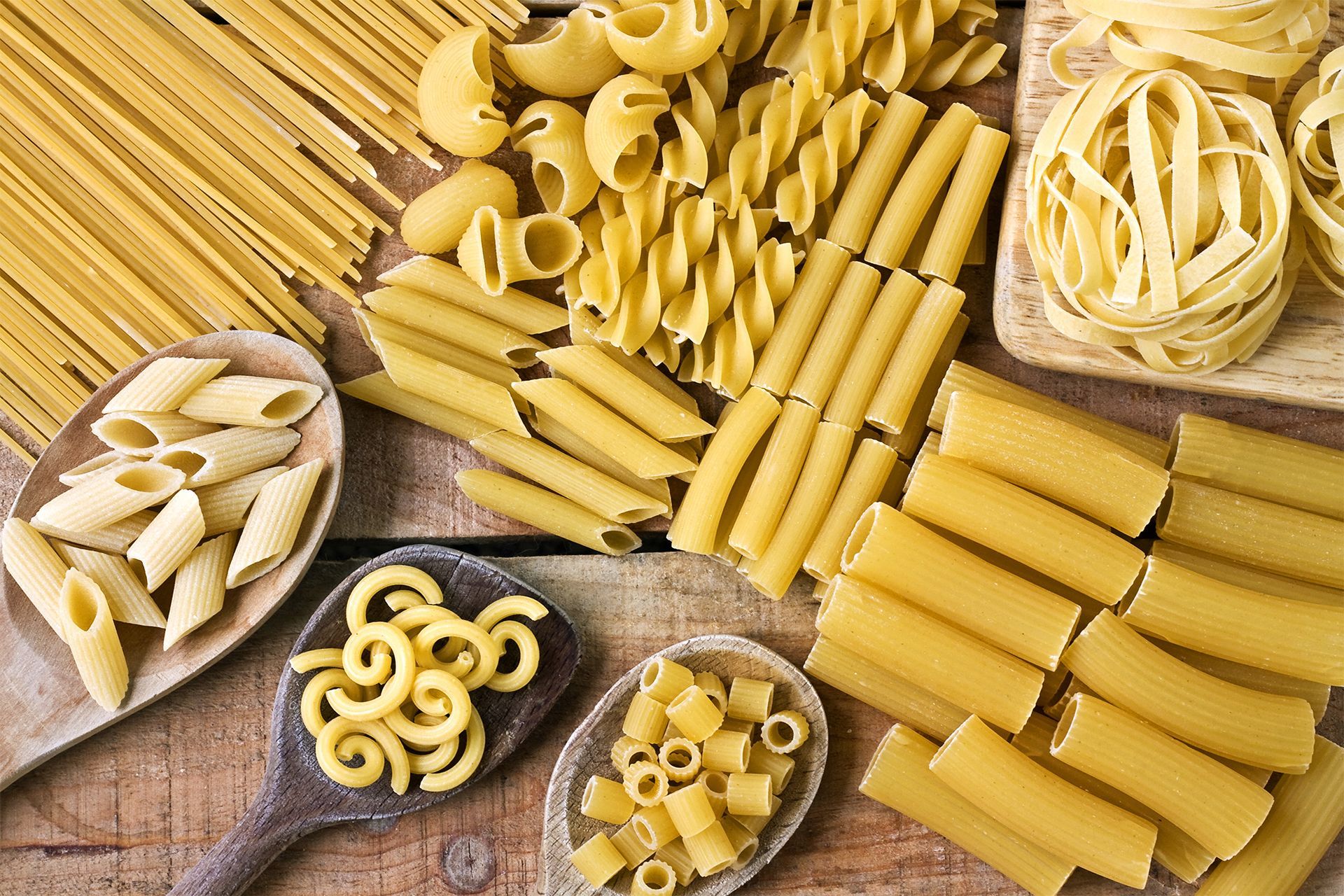 Assorted dried pasta shapes in bowls and scattered on a rustic wooden surface.