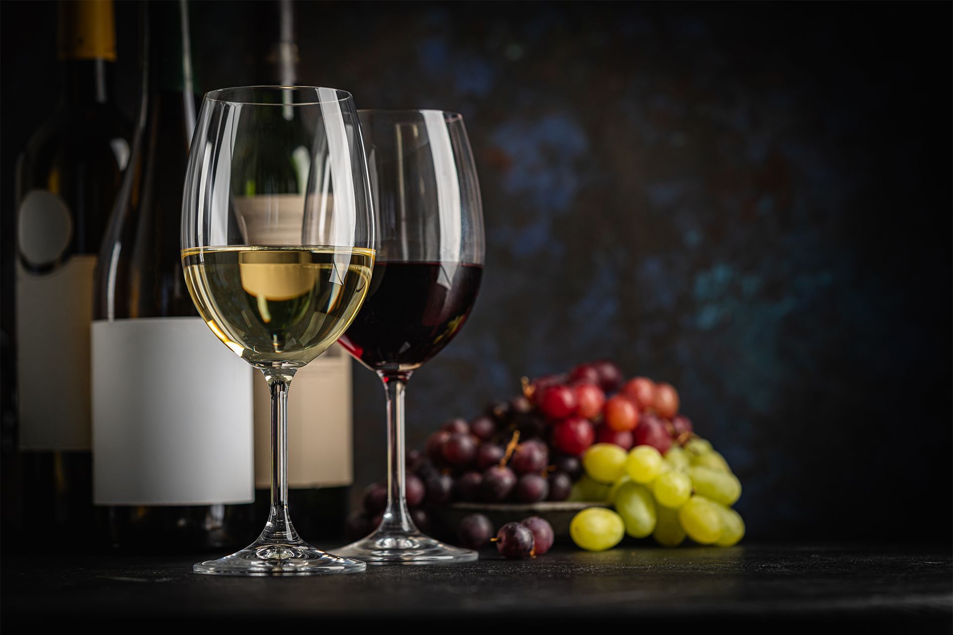 Wine glasses with red and white wine beside bottles and clusters of grapes on a dark table.