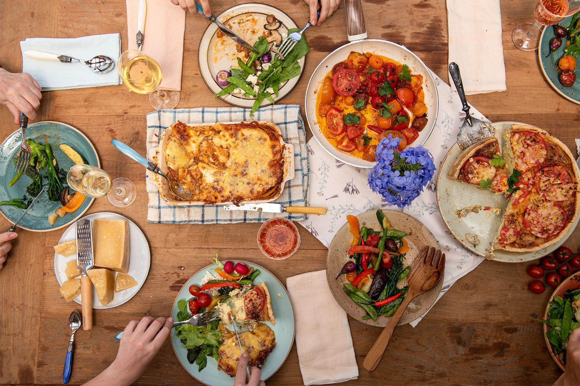 Overhead view of a rustic table with assorted colorful dishes, drinks, and hands serving food. Overhead view of a rustic table with assorted colorful dishes, drinks, and hands serving food.