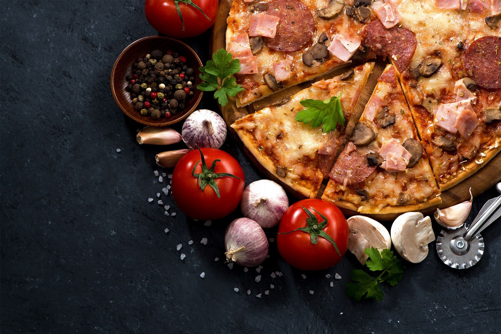 Top-down view of a sliced pizza with tomatoes, garlic, and spices on a dark table. Top-down view of a sliced pizza with tomatoes, garlic, and spices on a dark table.