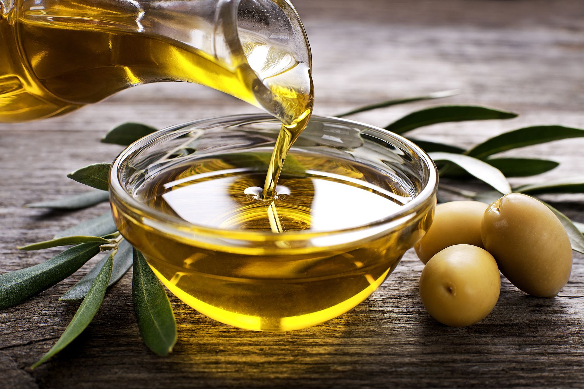 Golden olive oil pouring into a glass bowl with olives and olive leaves on a wooden surface. Golden olive oil pouring into a glass bowl with olives and olive leaves on a wooden surface.