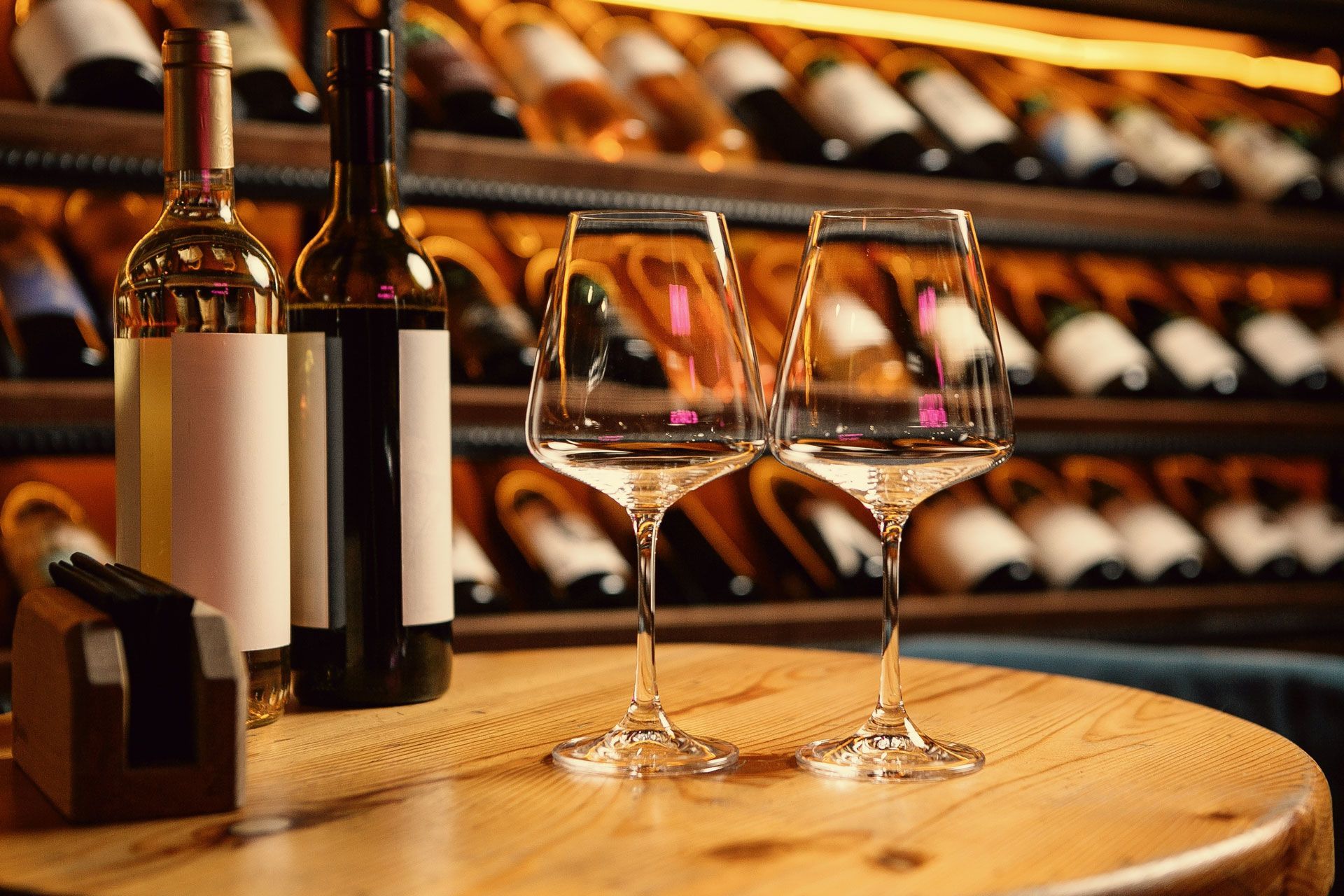 Two wine bottles and two glasses on a wooden table in front of a wine rack