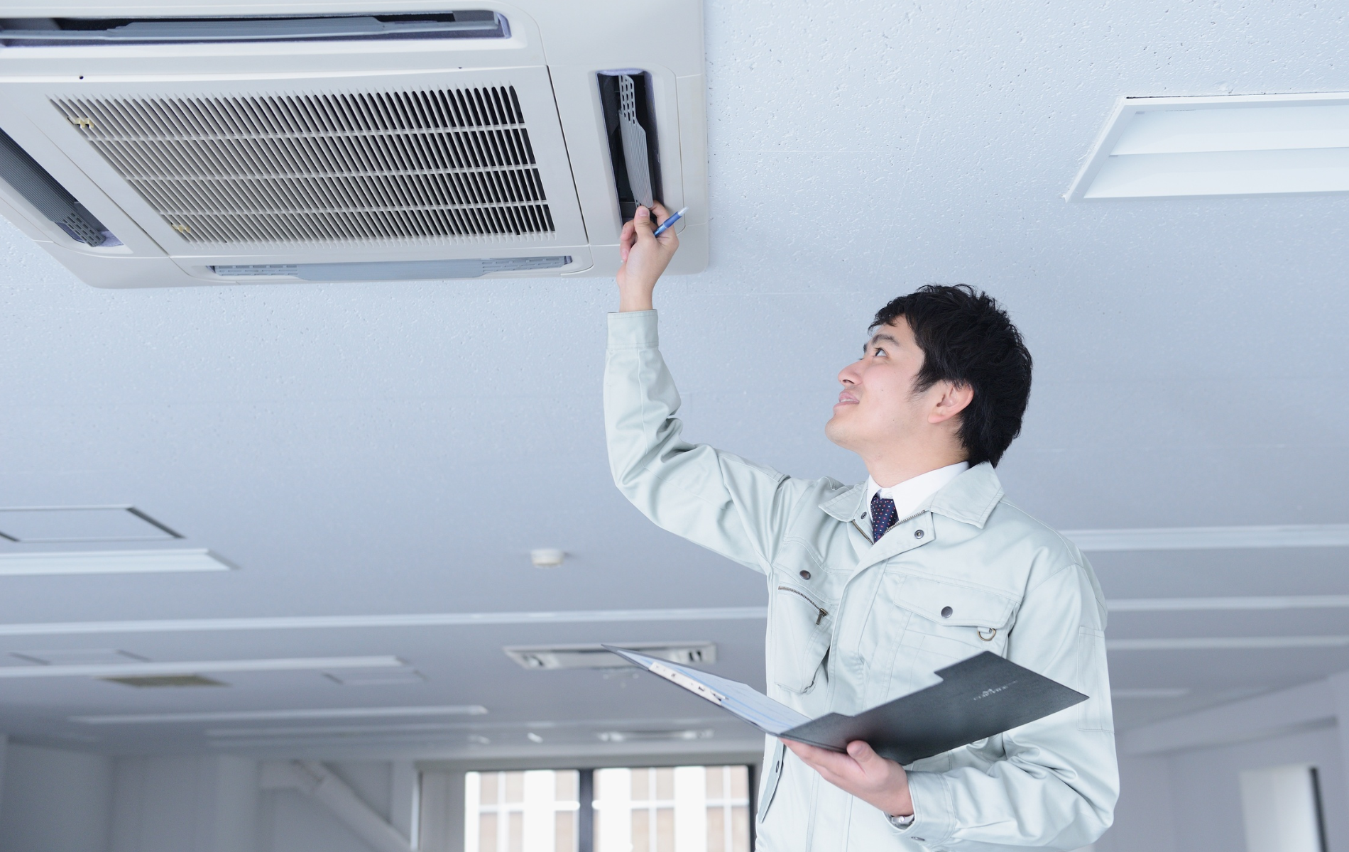 Man in work uniform inspecting an air conditioning unit in a building, holding a clipboard.