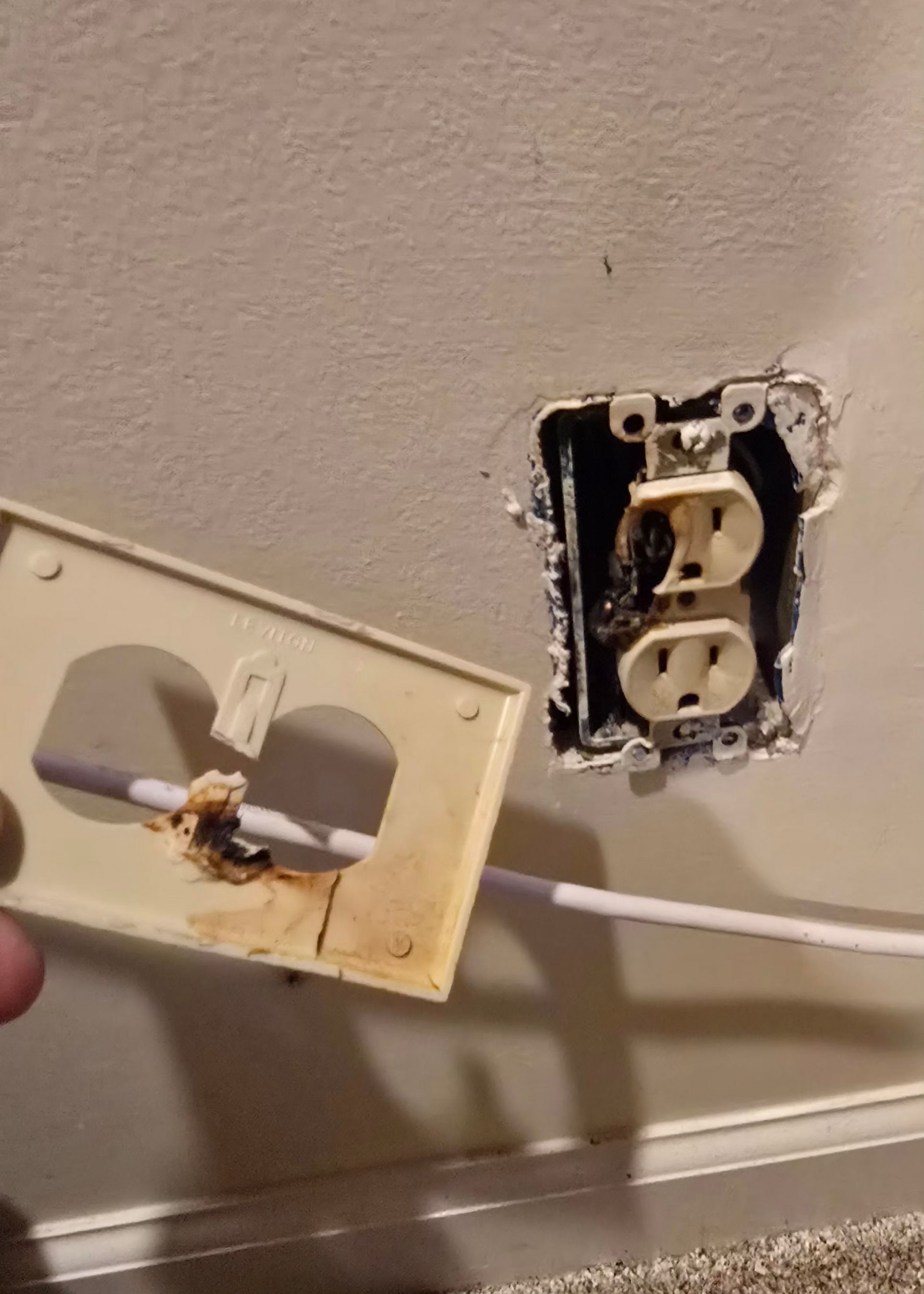 Damaged wall outlet with exposed wiring and a detached outlet cover plate held beside it