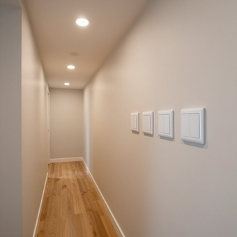 Narrow beige hallway with recessed ceiling lights, wooden floor, and white light switches on the right wall